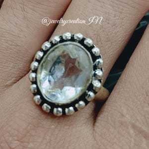 Può includere: Anello d'argento ovale con una grande pietra preziosa trasparente. L'anello ha un bordo di perline attorno alla gemma. L'anello è su una mano con lo smalto rosa. Il testo "@jewelrycreation_JN" è visibile.