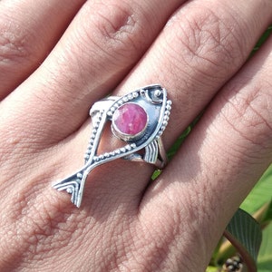 May include: A silver ring with a pink gemstone in the shape of a fish. The ring is on a person's finger.