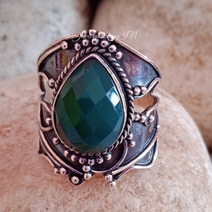 May include: A silver ring with a green gemstone set in a detailed, ornate design. The ring is on a background of smooth, light brown stones.