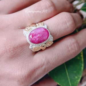Op de afbeelding: Een zilveren ring met gouden accenten en een grote, ovale roze edelsteen. De ring is geplaatst tegen een achtergrond van groene bladeren.