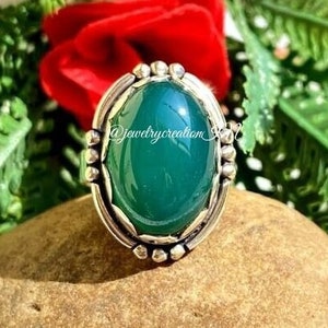 Könnte beinhalten: Ein Silberring mit einem großen, ovalen, smaragdgrünen Stein. Der Stein ist in einer dekorativen Silberfassung mit kleinen Perlenakzenten gefasst. Der Ring wird auf einem braunen Stein präsentiert, umgeben von grünem Laub und einer roten Rose.