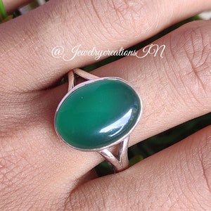 May include: A silver ring with a green oval gemstone. The ring is on a finger and the background is a green plant.