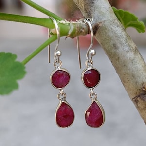 以下が含まれることがあります： 2 つの赤い宝石の雫が付いたシルバーのドロップイヤリングのペア。 上部の宝石は丸く、下部の宝石は涙型です。