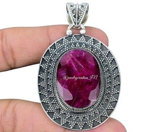 May include: A silver pendant with a large, oval-shaped, pink ruby gemstone set in a silver frame with a decorative border. The pendant is on a finger.