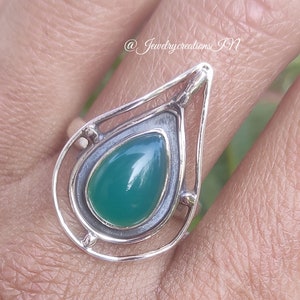 May include: A silver ring with a teardrop-shaped green gemstone set in a delicate, geometric design. The ring is worn on a person's finger.
