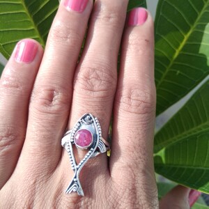 May include: A silver ring with a pink gemstone in the shape of a fish. The ring is on a person's finger.