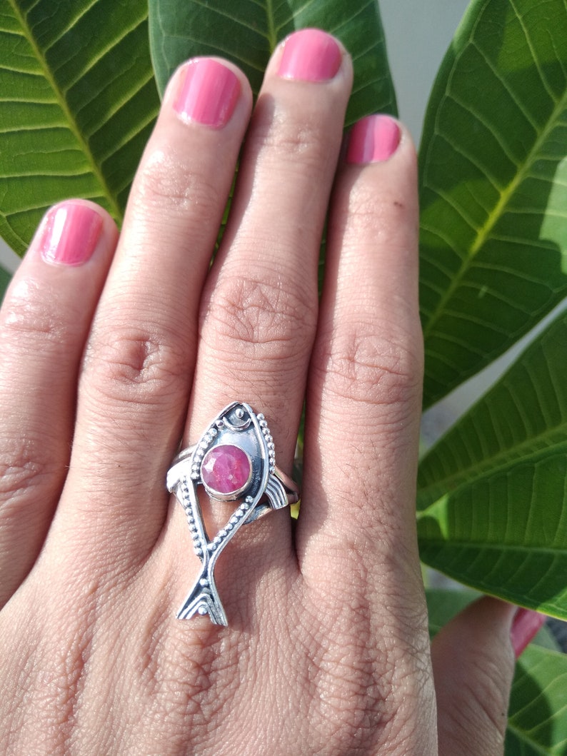 May include: A silver ring with a pink gemstone in the shape of a fish. The ring is on a person's finger.