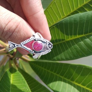 May include: A silver ring with a red gemstone in the shape of a fish. The ring is on a finger and the background is a green leaf.
