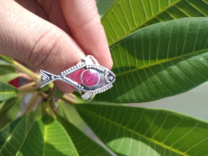 May include: A silver ring with a red gemstone in the shape of a fish. The ring is on a finger and the background is a green leaf.
