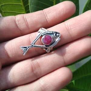 May include: A silver ring with a pink gemstone set in the center of a fish design. The ring is on a finger.