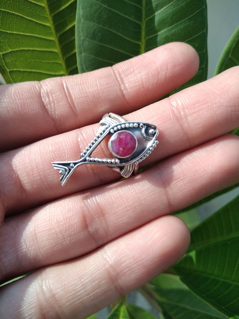May include: A silver ring with a pink gemstone set in the center of a fish design. The ring is on a finger.