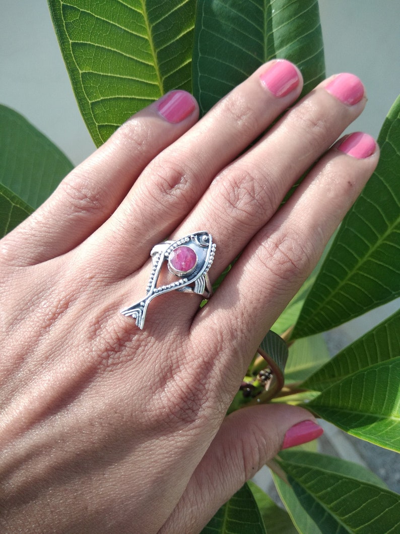 May include: A silver ring with a pink gemstone in the shape of a fish. The ring is on a person's finger.