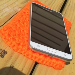 Puede incluir: Un teléfono inteligente Samsung blanco con una pantalla negra descansa sobre una funda de teléfono de ganchillo de color naranja brillante.