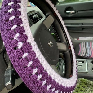 May include: Purple and white crocheted steering wheel cover. The cover is made with a simple stitch pattern and has a white stripe around the center.