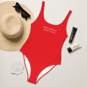 May include: Red one-piece swimsuit with white straps and a white trim. The swimsuit has the text "Make America Great Again." printed on the front.