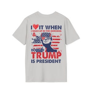 Pode incluir: Uma camiseta cinza com um coração vermelho, as palavras "I ♥ IT WHEN I WAKE UP IN THE MORNING & DONALD TRUMP IS PRESIDENT", e um gráfico de Donald Trump com um fundo de bandeira americana.