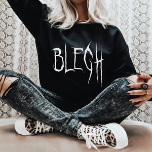 以下が含まれることがあります： 前面に白い文字で「BLEGH」とプリントされた黒いスウェットシャツ。スウェットシャツは、足を組んで座っている人が着用しており、破れたジーンズとヒョウ柄のスニーカーを履いています。