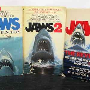 May include: Three paperback books featuring Jaws, Jaws 2, and Jaws The Revenge. Each cover displays a shark emerging from blue water with white text titles and author names. The books are arranged side-by-side, showing illustrations of sharks.