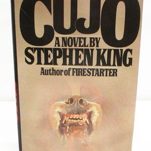 May include: A vintage paperback book titled "CUJO" by Stephen King. The cover features the title in large black letters, the author's name, and a menacing illustration of a dog's snarling face with exposed teeth and saliva.