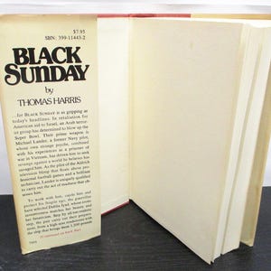 May include: A vintage hardcover book, "Black Sunday" by Thomas Harris, with a cream-coloured cover and black lettering. The book is open, displaying the text and pages. The spine is red.