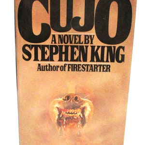 May include: A vintage paperback book titled "CUJO" by Stephen King. The cover features the title in large, dark brown letters, with the author's name and a menacing dog illustration.