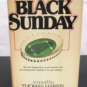 May include: A vintage paperback book titled "BLACK SUNDAY" by Thomas Harris. The cover shows a stadium illustration with a green field. The text reads, "Do not begin this novel unless you are prepared to finish it in one sitting..."