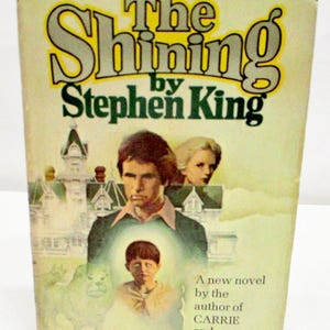 May include: A hardcover book titled The Shining by Stephen King with a pale yellow cover. The illustration features a man, a woman, and a child in front of a large house. Text on the cover includes A new novel by the author of CARRIE and SALEM'S LOT.