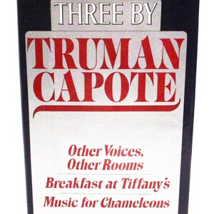 May include: A book cover for "Three by Truman Capote" with the author's name in large red lettering. The cover lists three of Capote's works: "Other Voices, Other Rooms," "Breakfast at Tiffany's," and "Music for Chameleons."