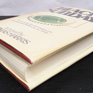May include: A hardback book titled "Black Sunday" by Thomas Harris. The book has a cream-coloured cover with a football field graphic and red edges. The book is lying on a black surface.
