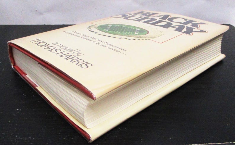 May include: A hardback book titled "Black Sunday" by Thomas Harris. The book has a cream-coloured cover with a football field graphic and red edges. The book is lying on a black surface.