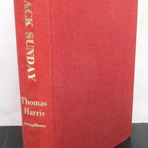 May include: A red hardcover book titled "Black Sunday" by Thomas Harris. The book's spine features the title and author's name in gold lettering. The cover is a solid red colour.