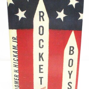 May include: Book cover for "Rocket Boys" by Homer H. Hickam, Jr. The design features a red and blue background with white stars and rocket-shaped text. The title "Rocket Boys" is prominently displayed.