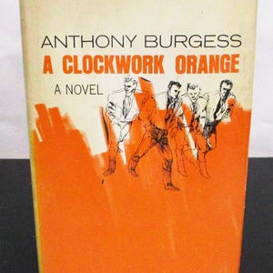 May include: A vintage paperback book titled "A Clockwork Orange" by Anthony Burgess. The cover features an orange and cream color scheme with illustrations of figures in suits. The book is a novel.