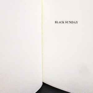 May include: An open book with the title "BLACK SUNDAY" printed in a dark brown font on the right page. The left page is blank. The pages are off-white with a textured surface.