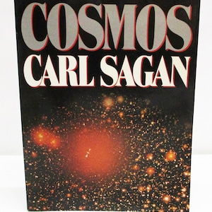 May include: A book cover with the title "COSMOS" in large, silver letters with red outlines, above the author's name, "CARL SAGAN," in white. The background is a dark space scene with a bright orange nebula and numerous stars.