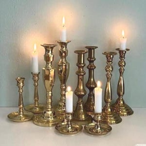 May include: A collection of brass candlesticks of varying heights and designs, some with lit white candles. The candlesticks have a polished, reflective surface, and are arranged on a white surface against a neutral background. The flames cast a warm glow.
