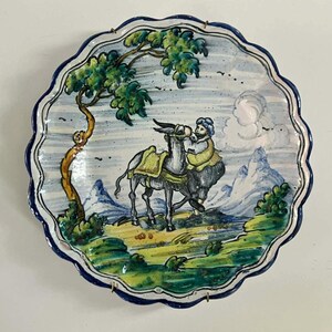 May include: A round ceramic plate with a scalloped edge featuring a hand-painted scene of a person standing beside a donkey. The landscape includes a tree, rolling green hills, and distant mountains under a sky with clouds, all in blue, green, and yellow.