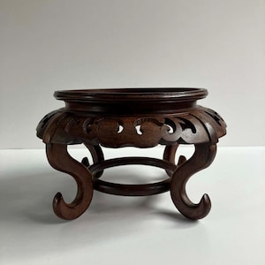 May include: A dark brown wooden display stand featuring a circular top with a raised rim, an ornate carved apron with openwork detailing, and four curved legs that connect to a central circular support ring at the base of the structure.