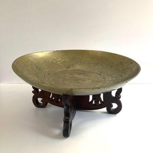 May include: A vintage brass bowl with intricate etched designs, resting on a dark wooden stand with carved details. The bowl has a wide, shallow shape and a golden hue. The stand has a tripod base.