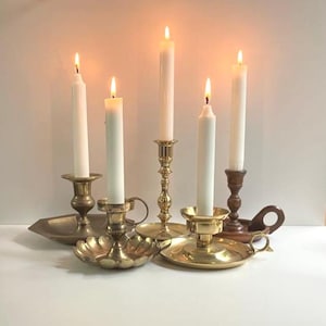 May include: Five lit white candles in various brass and wooden candle holders. The candle holders have different designs, including ornate, fluted, and handled styles. The scene is set against a neutral background, with soft lighting.