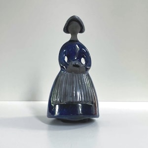 May include: A ceramic figurine of a woman in a blue dress and bonnet. The figure has a glossy finish and is holding a basket. The dress has vertical lines, and the figure has a simple, stylized design. The figurine is on a white background.
