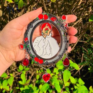 Puede incluir: Un pin de metal plateado con un dise&ntilde;o de esmalte rojo y blanco. El pin presenta un retrato de una persona con un coraz&oacute;n rojo que le cubre el ojo. El pin est&aacute; rodeado de una corona de rosas rojas y tiene tres colgantes de coraz&oacute;n rojo.