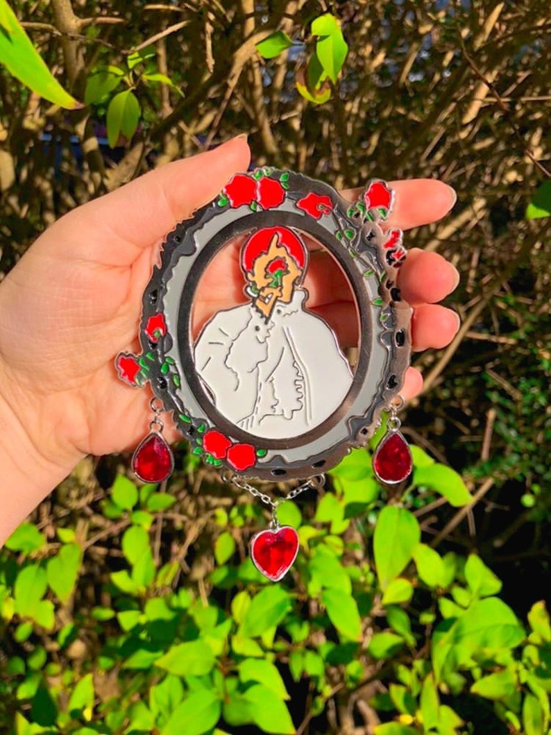 Puede incluir: Un pin de metal plateado con un dise&ntilde;o de esmalte rojo y blanco. El pin presenta un retrato de una persona con un coraz&oacute;n rojo que le cubre el ojo. El pin est&aacute; rodeado de una corona de rosas rojas y tiene tres colgantes de coraz&oacute;n rojo.
