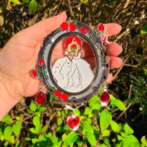 Puede incluir: Un pin de metal plateado con un retrato rojo y blanco dentro de un marco circular. El marco est&aacute; decorado con rosas rojas y hojas verdes. Tres colgantes rojos en forma de coraz&oacute;n cuelgan de la parte inferior del marco.