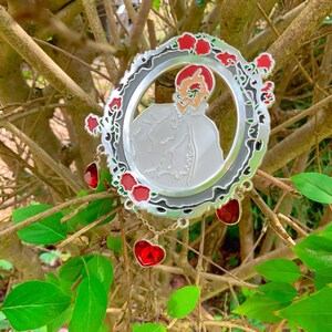 Puede incluir: Un pin de metal plateado con un dise&ntilde;o de esmalte rojo y blanco de una persona en un marco rodeado de rosas rojas. El pin tiene una cadena con tres dijes en forma de coraz&oacute;n rojo.