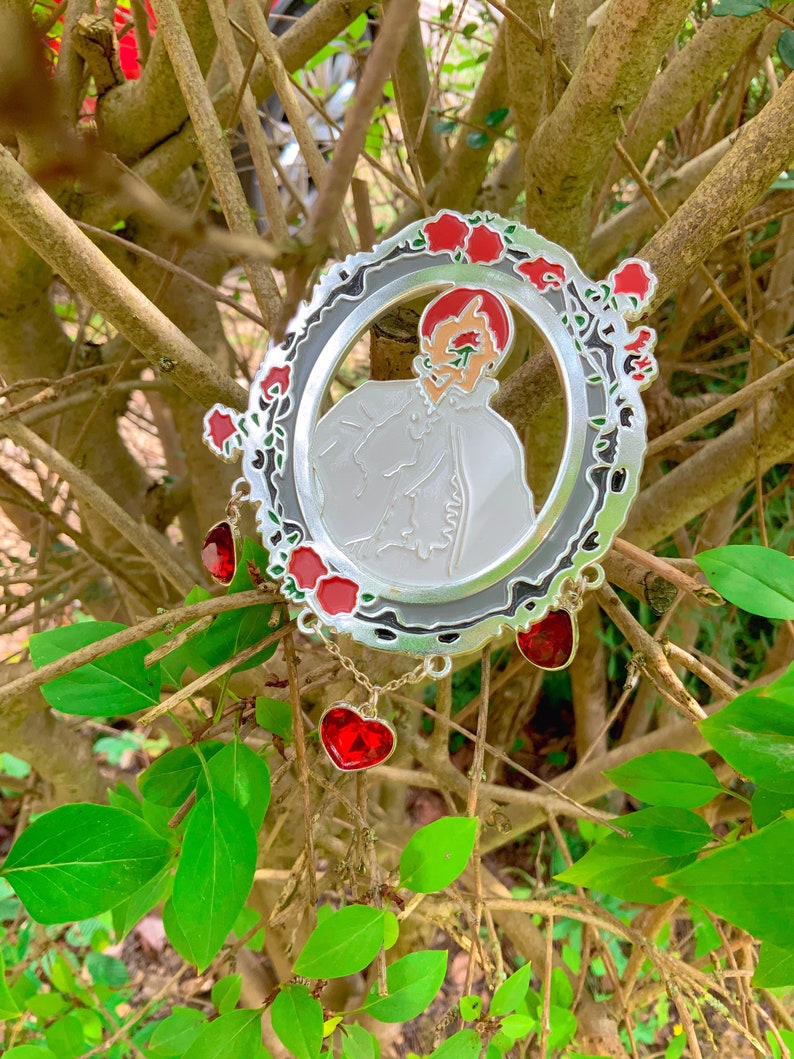 Puede incluir: Un pin de metal plateado con un dise&ntilde;o de esmalte rojo y blanco de una persona en un marco rodeado de rosas rojas. El pin tiene una cadena con tres dijes en forma de coraz&oacute;n rojo.