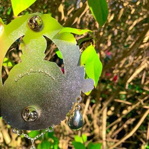 Puede incluir: Pin de metal plateado con un dise&ntilde;o detallado de una mujer con una corona y un colgante en forma de coraz&oacute;n. El pin tiene dos cierres en la parte posterior y un encanto de l&aacute;grima colgante.
