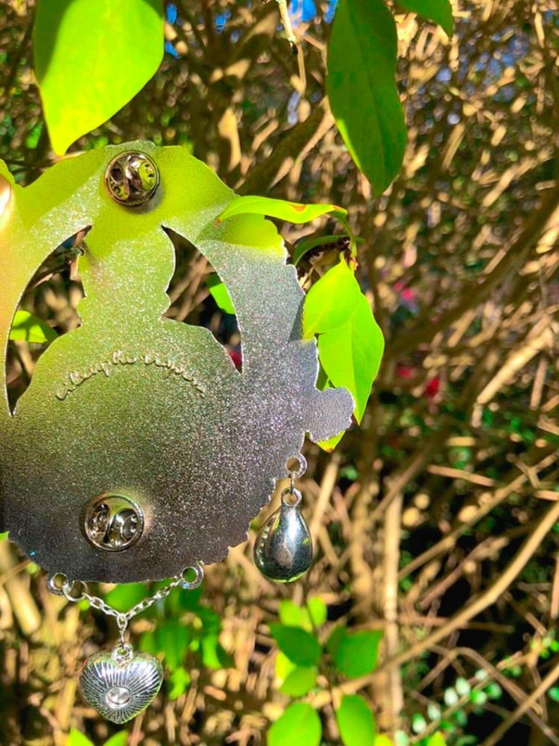 Puede incluir: Pin de metal plateado con un dise&ntilde;o detallado de una mujer con una corona y un colgante en forma de coraz&oacute;n. El pin tiene dos cierres en la parte posterior y un encanto de l&aacute;grima colgante.
