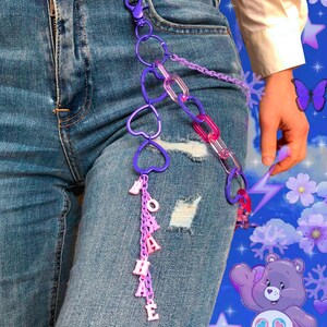 Puede incluir: Un accesorio de cadena de plástico morado y rosa sujeto a unos vaqueros azules. El accesorio presenta eslabones en forma de corazón, eslabones de cadena translúcidos y una hilera vertical de cuentas con letras rosas que forman BOHAE.