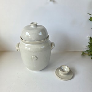 May include: A light grey ceramic butter keeper with a rounded lid and two side handles. The main vessel features a small circular embossed shell detail on the front. A separate small circular ceramic base piece sits to the right of the jar.